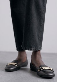 Osoba o ciemnej karnacji ubrana w krótkie czarne spodnie i czarne skórzane buty loafers z detalami złotych metalowych klamr, stojąca na szarym podłożu.