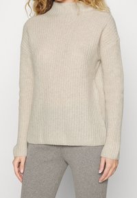 Femme portant un pull à col roulé côtelé beige et un pantalon côtelé gris, se tenant avec les bras détendus le long du corps.