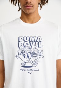 Balta marškinėliai su animaciniu personažu dubenyje su maistu, mėlyna tekstas „PUMA BOWL“ ir po juo „Mėgaukitės sveiku užkandžiu“. 