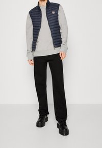 Navy puffer vest over a light grey sweatshirt, paired with black trousers and chunky black boots. The vest has a zip and logo detail.