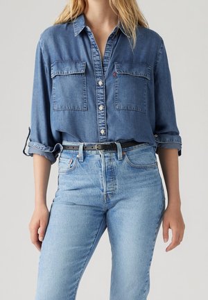 Femme portant une chemise en denim bleu avec manches retroussées et un jean bleu clair avec ceinture noire sur fond uni.