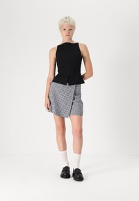 Čierny top bez rukávov s prednými gombíkmi, doplnený čierno-bielou suknou v štýle houndstooth. Model má na sebe čierne topánky a biele ponožky.