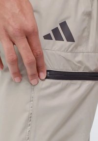 Beige träningsbyxor med en slät textur, som har en svart dragkedjeficka och en mörk logotyp bestående av tre ränder på låret.