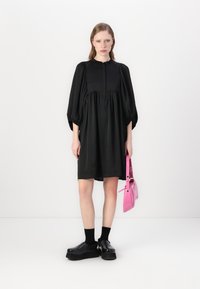 Vestido preto com mangas balão e detalhe de botões na frente, feito de tecido suave. Usado com sapatos pretos chunky e uma bolsa rosa.