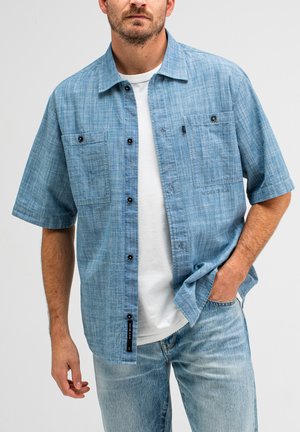 Korte mouwen blauwe knoopshirt van textuurstof, met twee borstzakken, zwarte knopen en een rechte zoom. Draag dit over een wit shirt.