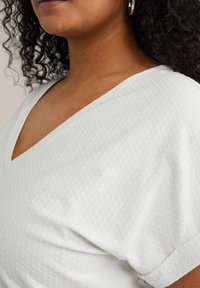 Top bianco testurizzato con scollatura a V e maniche corte, caratterizzato da un delicato motivo a pois e tessuto morbido.