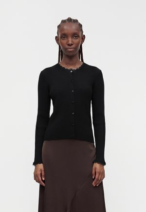 Fonott hajú nő fekete, gombos bordázott kardigánt és sötétbarna szoknyát visel egyszínű világos háttér előtt.