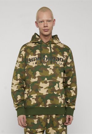 Person trägt grünen und braunen Tarnfleck-Hoodie und passende Hose mit Textlogo auf der Brust und der Hosentasche, steht vor schlichtem Hintergrund.