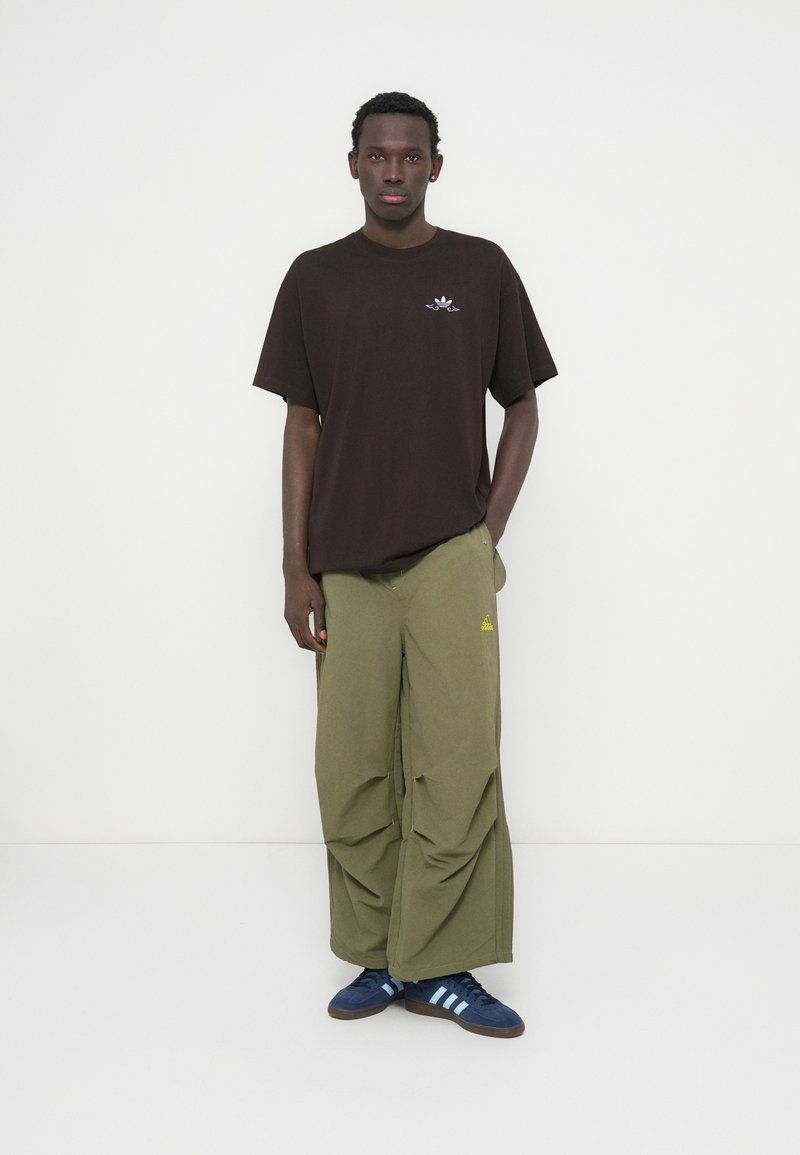 T-shirt marron surdimensionné avec un petit logo, pantalon ample vert olive avec des accents jaunes, et chaussures bleues avec des rayures blanches.