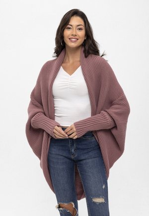 Strickjacke in sanftem Mauve, mit verlängerten Ärmeln und einem offenen Design. Kombiniert mit einem weißen Rippenshirt und zerrissenen blauen Jeans.