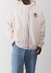 Sudadera con cremallera en color crema, con un logo frontal, bolsillos tipo canguro, puños acanalados y capucha forrada. Combinada con pantalones de mezclilla azul claro.