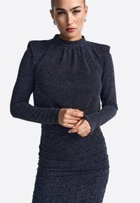 Vestido de color azul marino oscuro con mangas largas, cuello alto y tejido texturizado que presenta un sutil brillo. Detalles fruncidos en la cintura. Diseño ajustado.