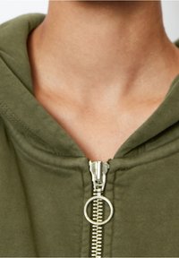 Felpa con zip color verde oliva, dotata di una cerniera metallica con un tirante circolare, texture morbida e un design del colletto rilassato.