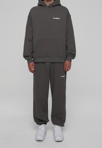 Grijze hoodie met een voorzak en een wit logo, gecombineerd met bijpassende grijze joggingbroek. Model draagt witte sneakers.