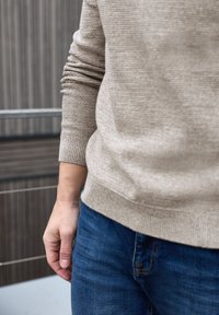 Beige neulepaita teksturoidulla kuviolla, resorit hihoissa ja helmassa. Yhdistetty sinisiin farkkuihin, jotka ilmaisevat rentoa istuvuutta ja muotoilua.