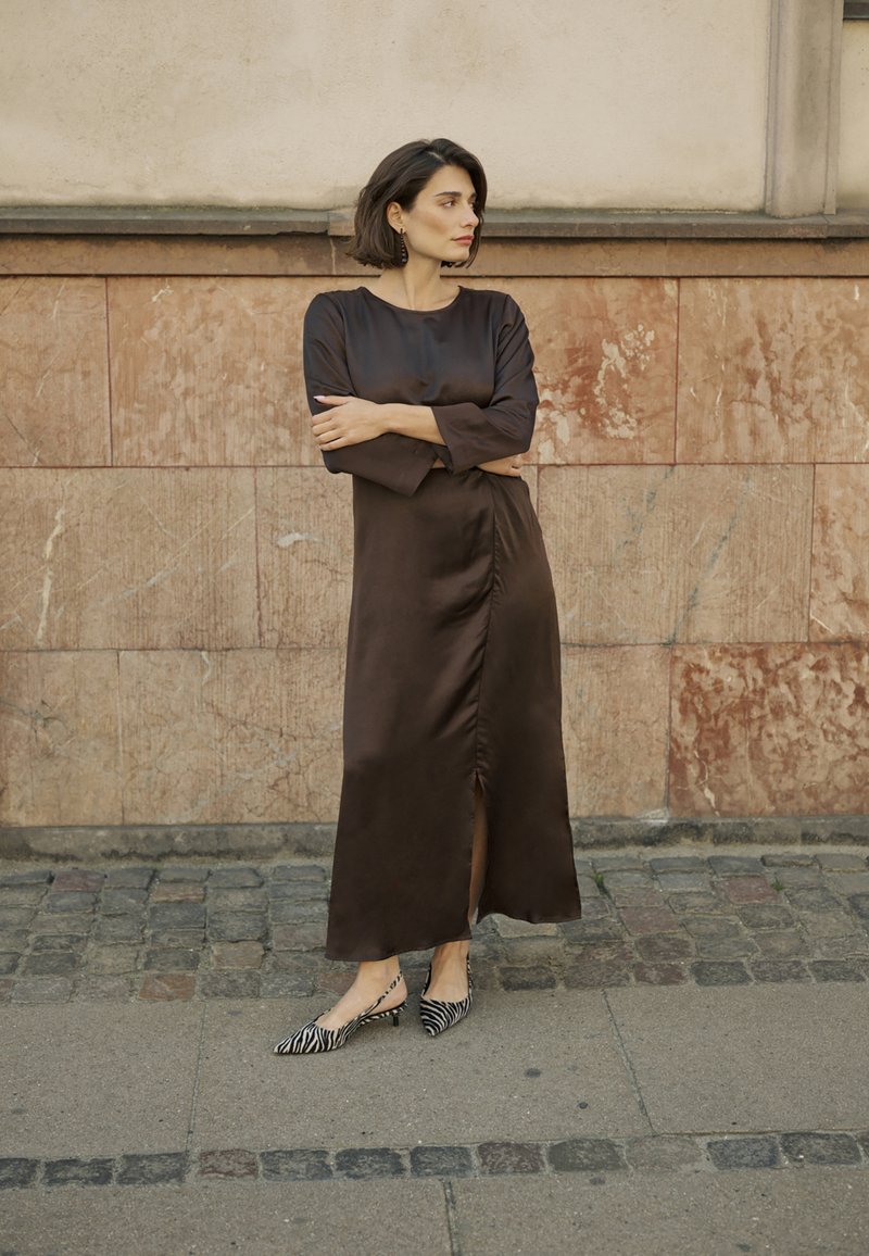 Brun satin klänning med långa ärmar, med en frontslits. Matchad med svarta och vita randiga spetsiga ballerina skor. Marmorvägg som bakgrund.