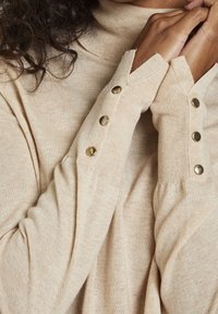 Beige-coloured poolopaita, jossa on ribbitekstuuria ja napituskoristeet hihoissa, rento istuvuus ja peukalon aukot.