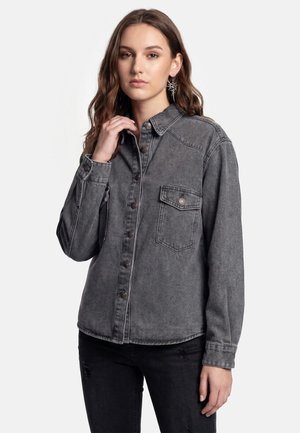 Kvinde med langt brunt hår iført grå cowboyjakke med knapper og sorte revnede jeans, der rører ved kraven med venstre hånd, neutral udtryk.