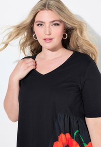 Blonde Frau mit Ohrhängern trägt ein schwarzes V-Ausschnitt-Shirt mit rotem Blumenmuster, hält den Kragen und hat weiches Haar, das vor einem weißen Hintergrund weht.