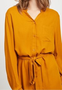 Chemise boutonnée jaune moutarde avec un décolleté en V, des manches longues, une poche sur la poitrine et une ceinture nouée à la taille. Tissu doux avec une texture lisse.
