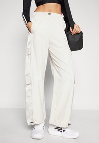 Lehké, krémové cargo kalhoty s elastickým pasem, bočními kapsami a zipovými akcenty, doplněné o bílé sportovní boty.