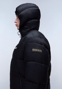 Chaqueta acolchada negra con capucha, que presenta un parche con el logo en la manga y un bolsillo con cremallera. Material texturizado y brillante, diseño acolchado.