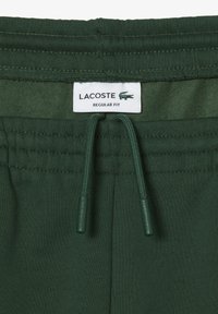 Pantalones de chándal verdes con una cinturilla elástica, cierre con cordón y una etiqueta que muestra "LACOSTE REGULAR FIT". Textura de tela suave.