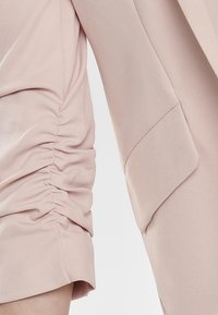 Light pink garment featuring gathered fabric detail on one sleeve and a smooth, tailored finish on the other. Textured and soft material.