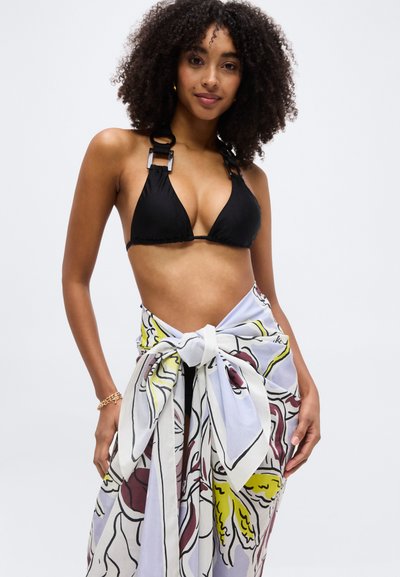 Γυναίκα με σγουρά μαλλιά που φοράει μαύρο μπικίνι τοπ με μεγάλα δαχτυλίδια και δεμένη φλοράλ σερφόνγκ, στέκεται μπροστά σε ένα απλό φωτεινό φόντο.