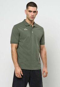 Mann med kort mørkt hår iført en olivengrønn poloshorte med en liten hvit logo på brystet og svarte bukser, stående foran en ensfarget vegg.