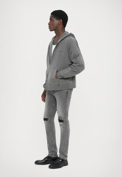 Grauer Reißverschluss-Hoodie mit Fronttasche, strukturierter Stoff. Getragen mit grauen, stark abgetragenen Jeans und schwarzen Schuhen, in seitlicher Position vor einem schlichten Hintergrund.
