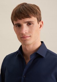 Junger Mann mit kurzem braunem Haar, trägt ein dunkelblaues Hemd mit Kragen, blickt nach vorne mit neutralem Gesichtsausdruck vor einem schlichten Hintergrund.