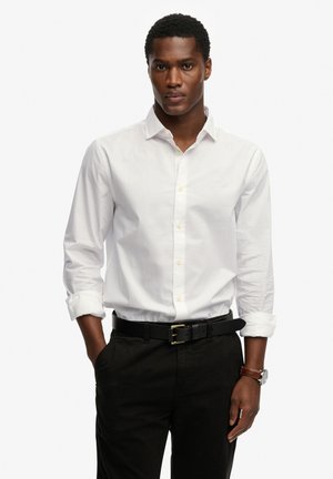 Weißes Baumwollhemd mit langen Ärmeln, durchgeknöpftem Vorderteil und leichtem Glanz, kombiniert mit schwarzen Hosen und einem schwarzen Gürtel mit goldener Schnalle.