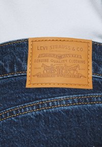 Denim materiale med en brun læderlap mærket "Levi Strauss & Co." med prægede tekster og heste grafik. Guld stikninger på sømmen.