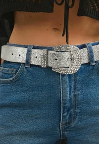 Sølvglitrende bælte med en stor, udsmykket spænde; struktureret overflade; båret over blå denimjeans.