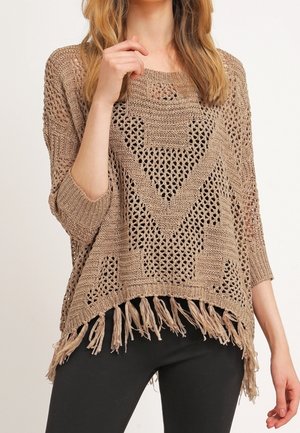 Femme portant un pull en maille crochet beige avec des motifs géométriques et un ourlet à franges, associée à un pantalon noir, debout devant un fond uni.