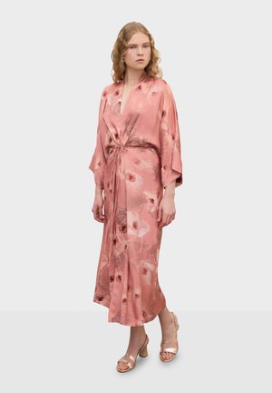 Žena oblečená v dlhej ružovej kvetinovej zavinovacej šaty so širokými rukávmi a viazaním v páse, v kombinácii s béžovými sandálmi na hrubom podpätku, stojaca na bielom pozadí.