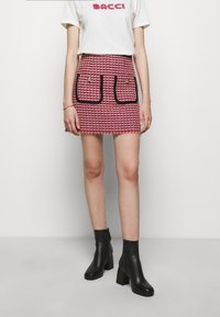 Mini-jupe à carreaux pied-de-poule rouge et blanc avec deux poches bordées de noir, terminée par une frange à l'ourlet. Associée à des bottines noires.