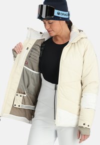 Puffjacke in hellem Beige mit weißen Akzenten, ausgestattet mit Kapuze, Frontreißverschluss und innerer Mesh-Tasche. Entworfen für Wärme und Wasserbeständigkeit.