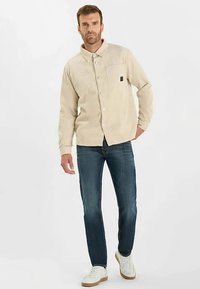Beige manchesterskjorta med knappar, två bröstfickor och ett logotypmärke, bärs med blå jeans och vita sneakers.