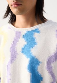 Sweter dziany w białym kolorze z zygzakowymi wzorami w odcieniach niebieskiego, purpurowego i żółtego. Posiada szeroki dekolt i fakturowany materiał. Luźny krój.