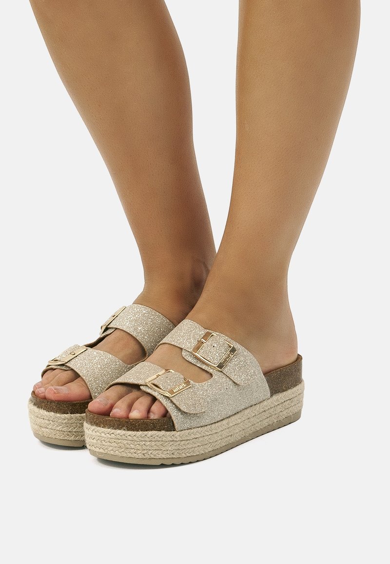 Pieds portant des sandales à plateforme beige pailletée avec des brides à double boucle et des semelles en corde tressée sur un fond clair.