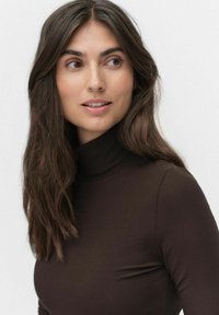 Brązowy, prążkowany top z golfem o dopasowanym kroju, miękkiej fakturze i wysokim kołnierzu, noszony przez osobę z długimi, ciemnymi włosami.