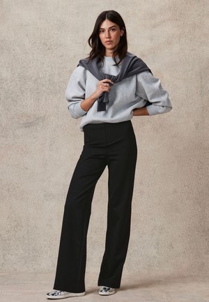 Femme debout contre un mur beige, portant un sweat gris, un pantalon noir taille haute à jambes larges, un pull gris posé sur les épaules et des baskets argentées.