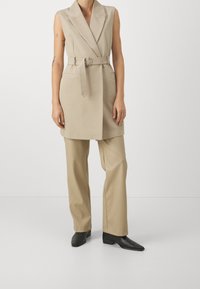 Beżowa kamizelka bez rękawów z paskiem w talii, ostrymi klapami i bocznymi kieszeniami, zestawiona z luźnymi beżowymi spodniami oraz czarnymi, spiczastymi butami.