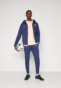 Navy tracksuit with a zippered hoodie and joggers, featuring light pink accents and a club logo. The person holds a soccer ball.