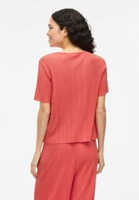 Top corto a maniche, di colore corallo, con una texture plissettata e una vestibilità rilassata, caratterizzato da un colletto rotondo e un orlo leggermente curvo.