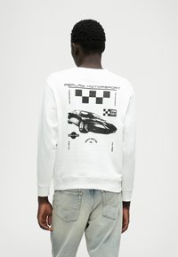 Balts džemperis ar melnu sporta automašīnas grafiku un šaha laukuma rakstu aizmugurē. Ribotās piedurknes un mala; kokvilnas maisījuma materiāls.