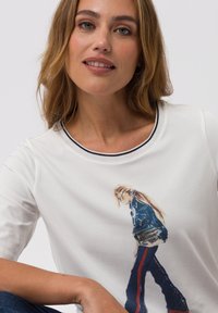 White cotton t-shirt featuring a printed design of a figure in a blue jacket and jeans. Rounded neckline with contrast stitching.