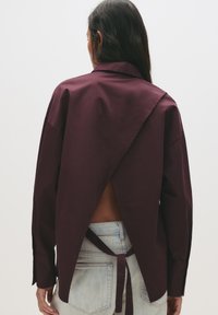 Femeie cu păr lung și închis la culoare, purtând o bluză maronie cu spatele liber legată la talie și blugi de denim de culoare albastră deschisă, întoarsă cu fața la un fundal simplu.
